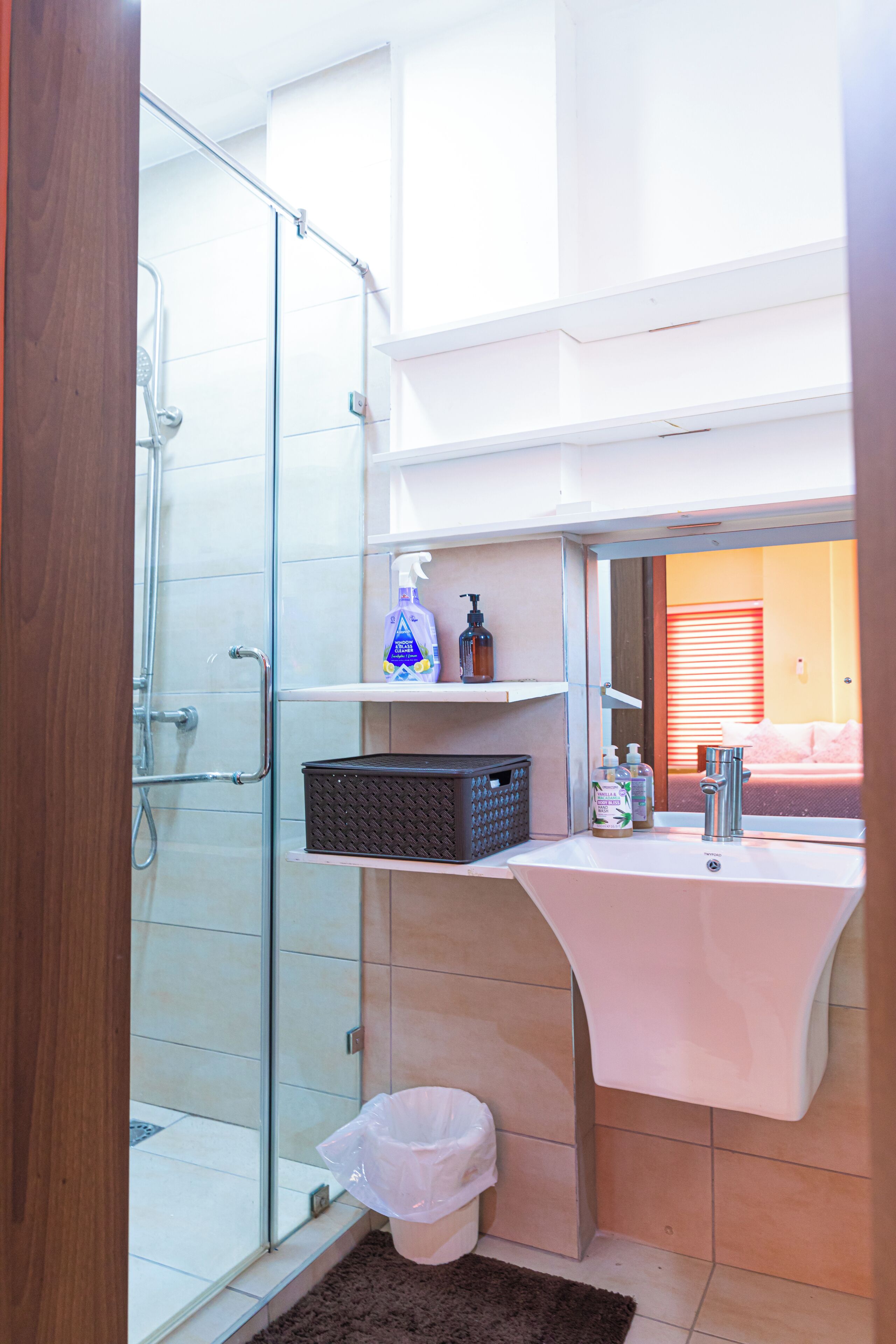 A bathroom with a glass shower enclosure, a white sink, and a mirror reflecting a room with a bed.