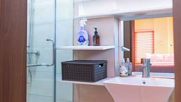 A bathroom with a glass shower enclosure, a white sink, and a mirror reflecting a room with a bed.