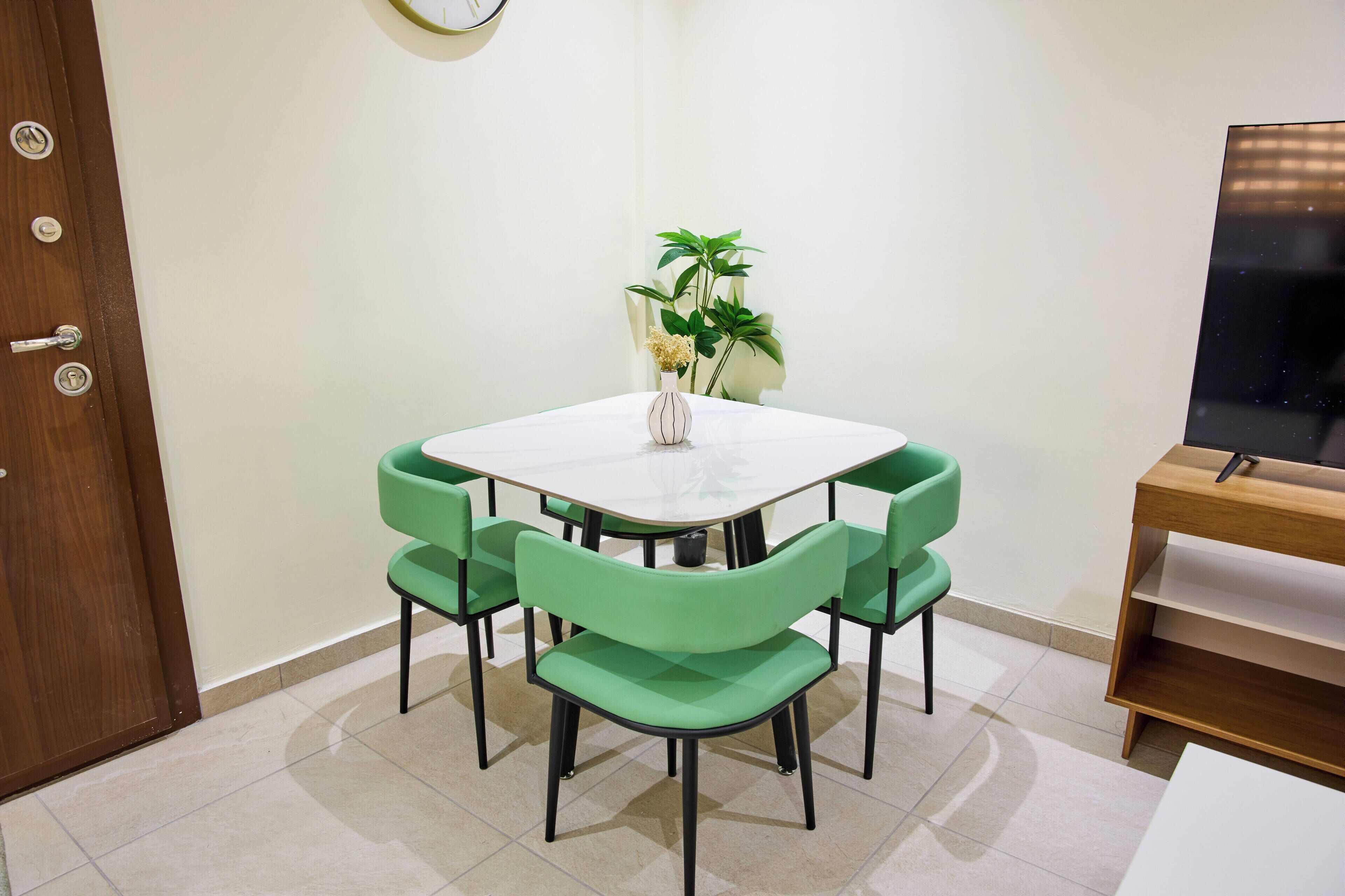A dining area with a round table, four green chairs, a vase with flowers, a plant, and a TV stand.