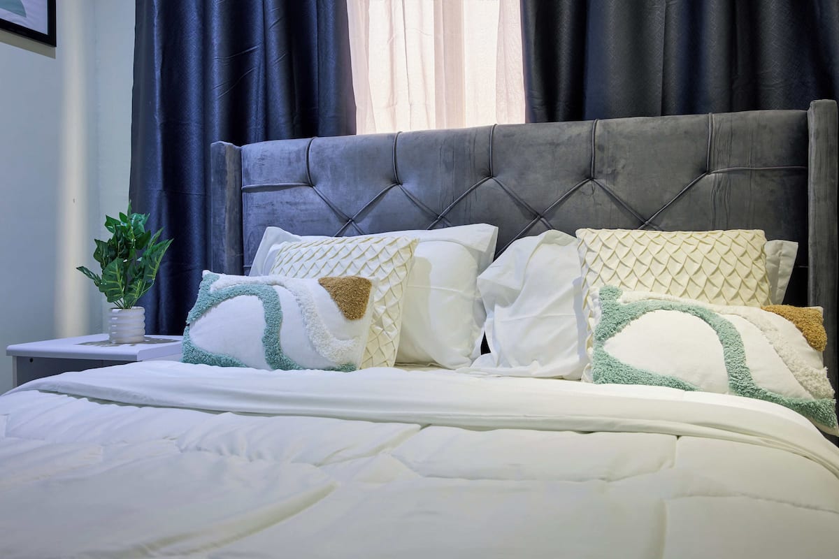 Una cama bien hecha con ropa de cama blanca y varios almohadones, una mesita de noche con una planta y una cortina oscura de fondo.