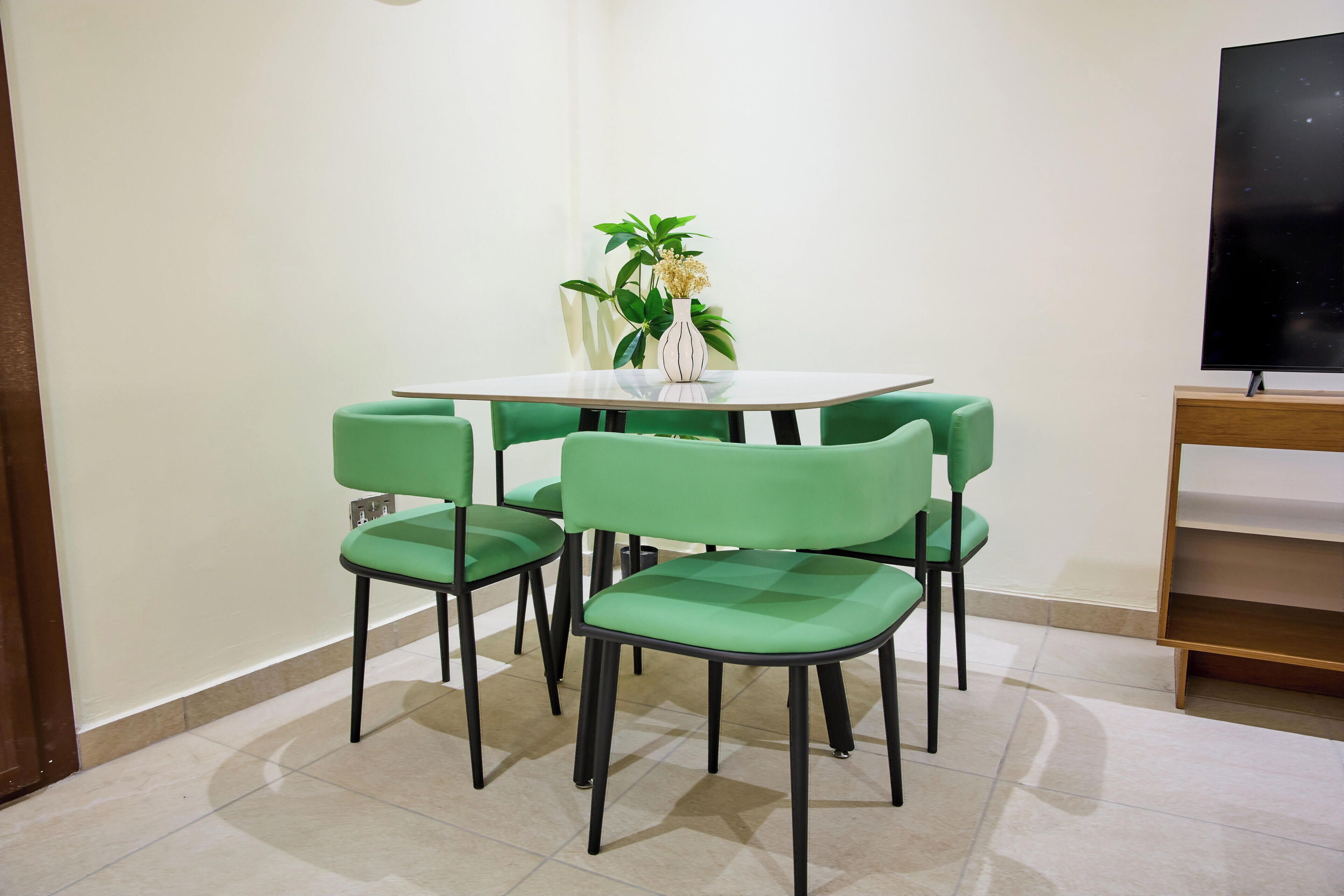 Un área de comedor con una mesa blanca y sillas verdes, un jarrón con flores y un televisor sobre un soporte de madera.