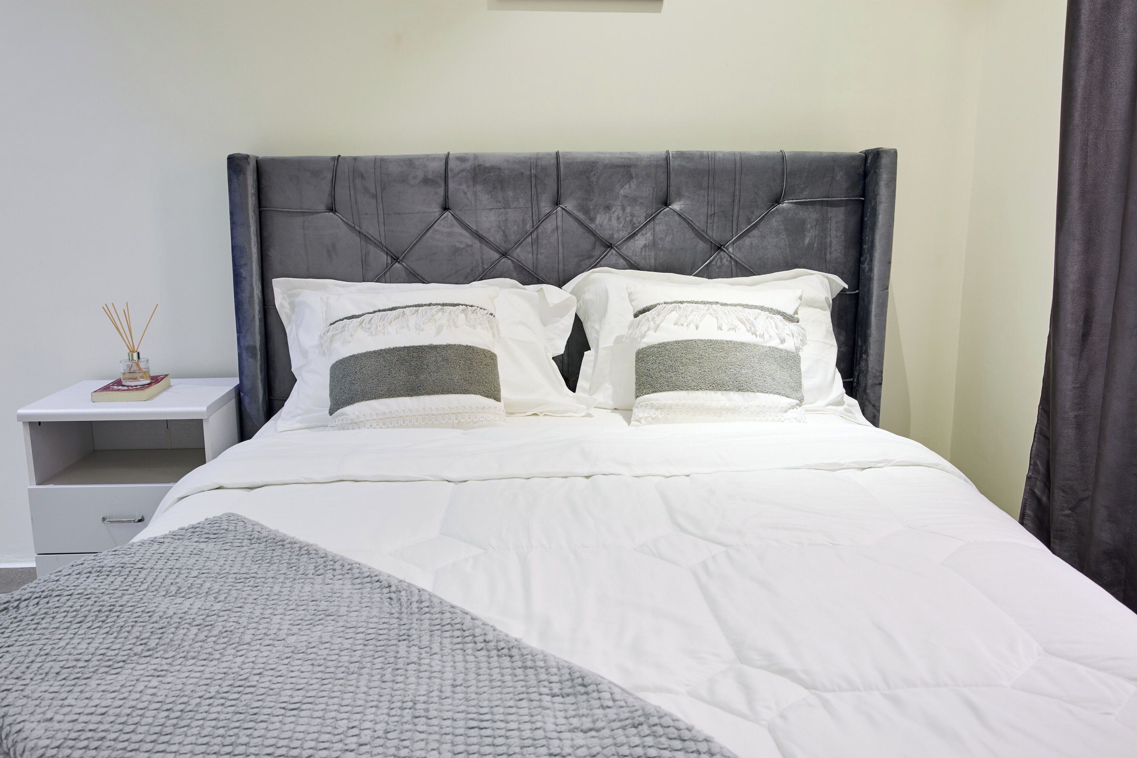 A neatly made bed with a grey upholstered headboard, white bedding, and a bedside table with a book and a reed diffuser.