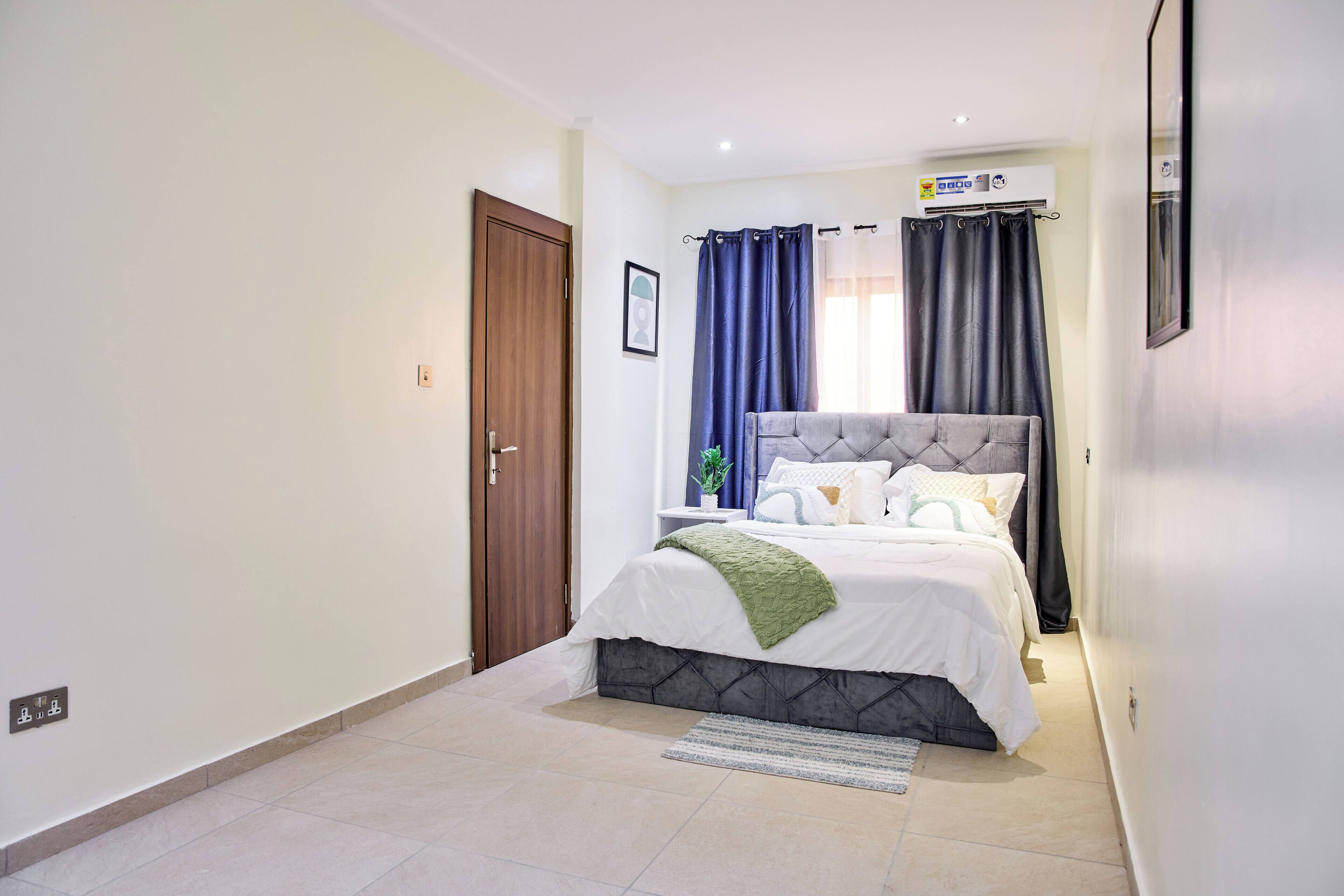 A bedroom with a bed, a door, a wall-mounted air conditioner, and a window with curtains.