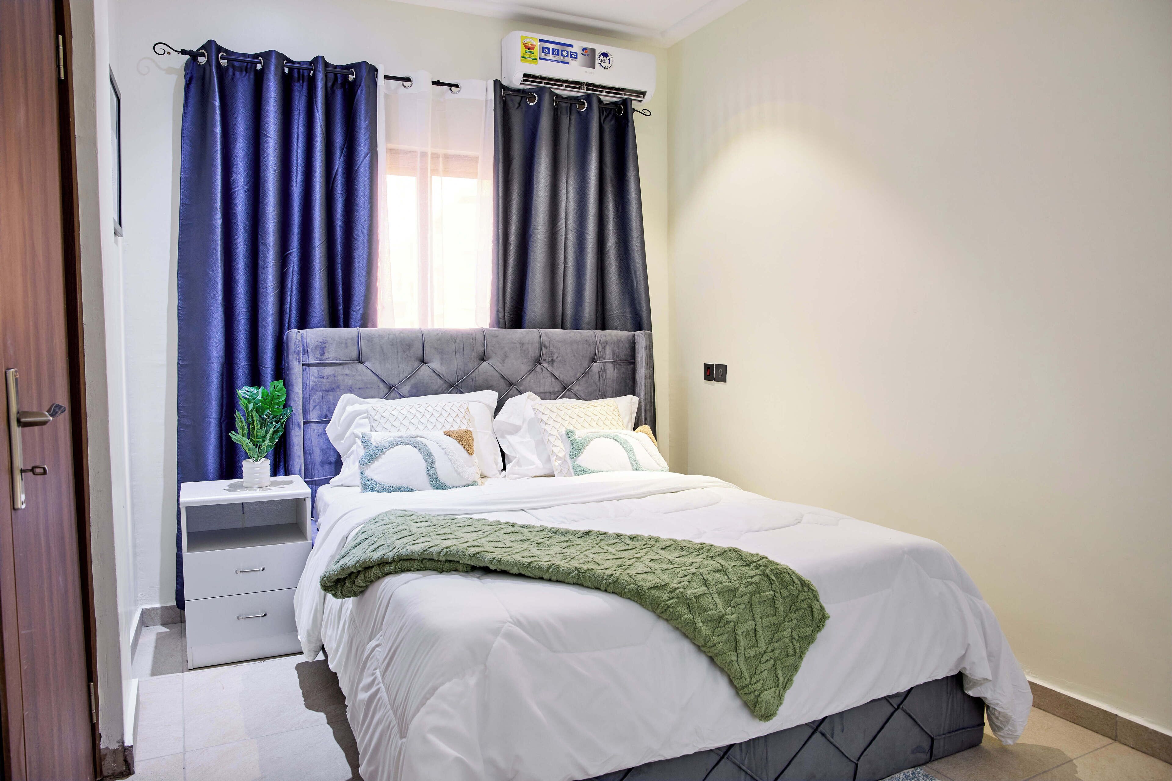 A neatly made bed with a grey headboard, a white comforter, and a green throw blanket. There is a bedside table with a plant and a lamp. The room has a window with blue curtains and an air conditioning unit above it.