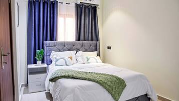 A neatly made bed with a grey headboard, a white comforter, and a green throw blanket. There is a bedside table with a plant and a lamp. The room has a window with blue curtains and an air conditioning unit above it.