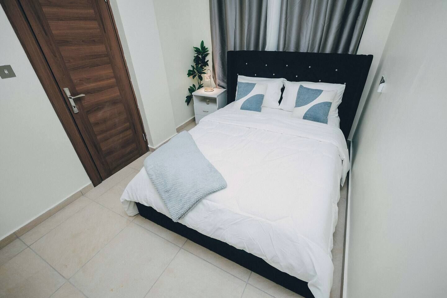 A neatly made bed with a white comforter and pillows, a bedside table with a lamp, and a door to the left.