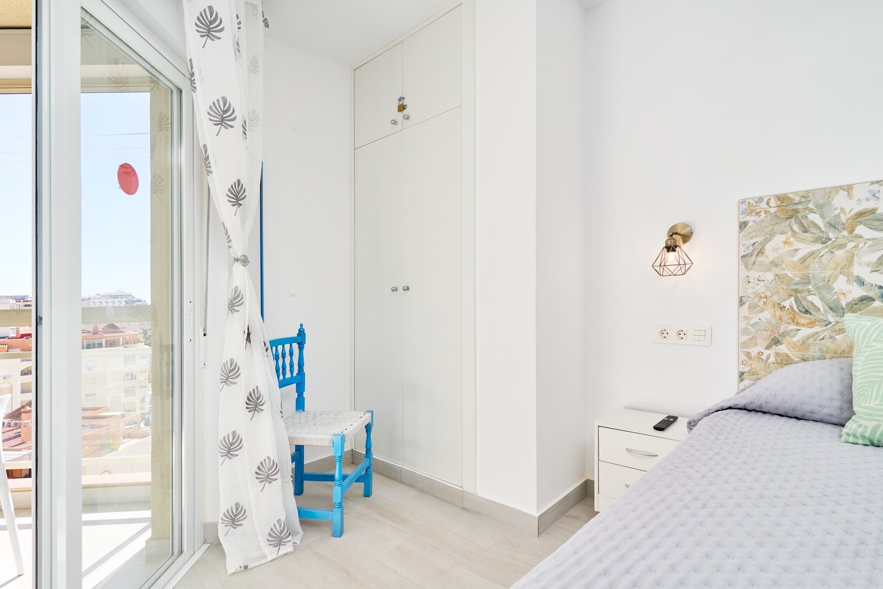 Un dormitorio con cama, una silla, una mesita de noche, una lámpara de pared y una ventana con cortina.