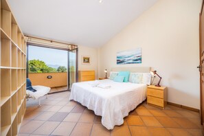 Une chambre à coucher comprenant un lit, des tables de chevet, une chaise, une porte-fenêtre coulissante donnant sur un espace extérieur et un tableau accroché au mur.