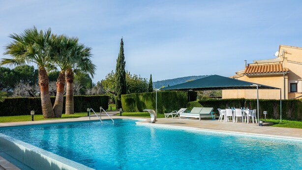 Ein Swimmingpool mit Liegestühlen und einem Tisch, umgeben von Palmen und einem Gebäude mit einem Ziegeldach.