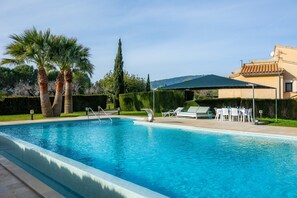 Ein Swimmingpool mit Liegestühlen und einem Tisch, umgeben von Palmen und einem Gebäude mit einem Ziegeldach.