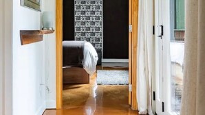 Un couloir avec un plancher en bois, un lit à l’arrière-plan, un miroir sur la gauche et une porte donnant sur une autre pièce.