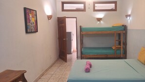 Una habitación pequeña y funcional con una litera, una cama individual con sábana verde, un banco de madera y un cuadro colgado en la pared.