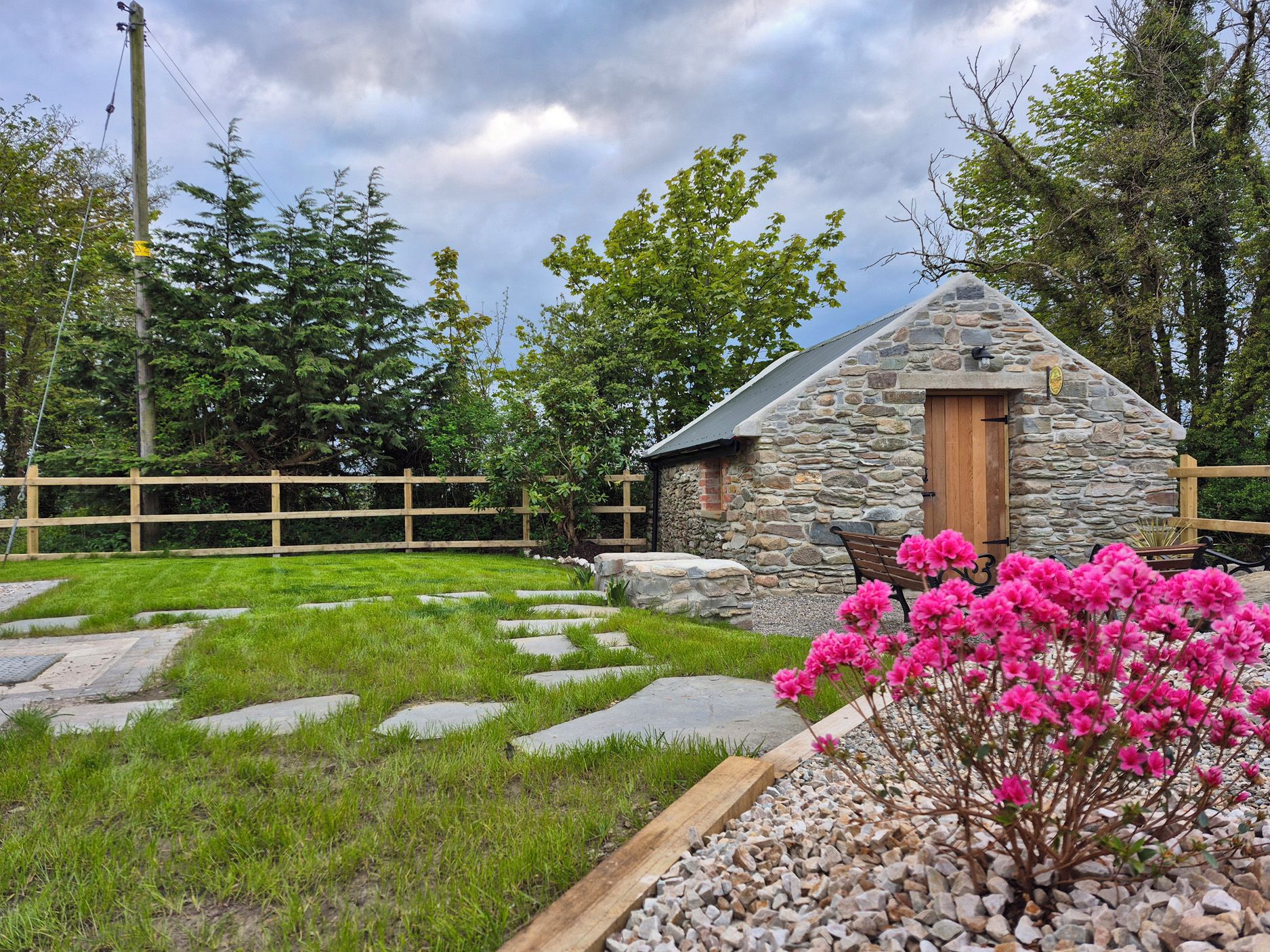 Una cabaña de piedra con puerta de madera, rodeada por un césped con piedras para pisar, una valla de madera y plantas con flores.