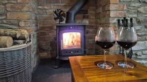 A cozy room with a brick wall, a wood-burning stove, a wicker basket with logs, and two wine glasses on a wooden table. - Annies Farm Cottage (Londonderry)