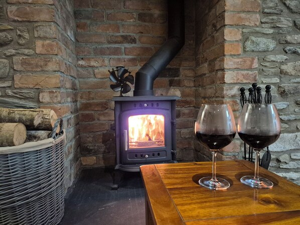 A cozy room with a brick wall, a wood-burning stove, a wicker basket with logs, and two wine glasses on a wooden table. - Annies Farm Cottage (Londonderry)