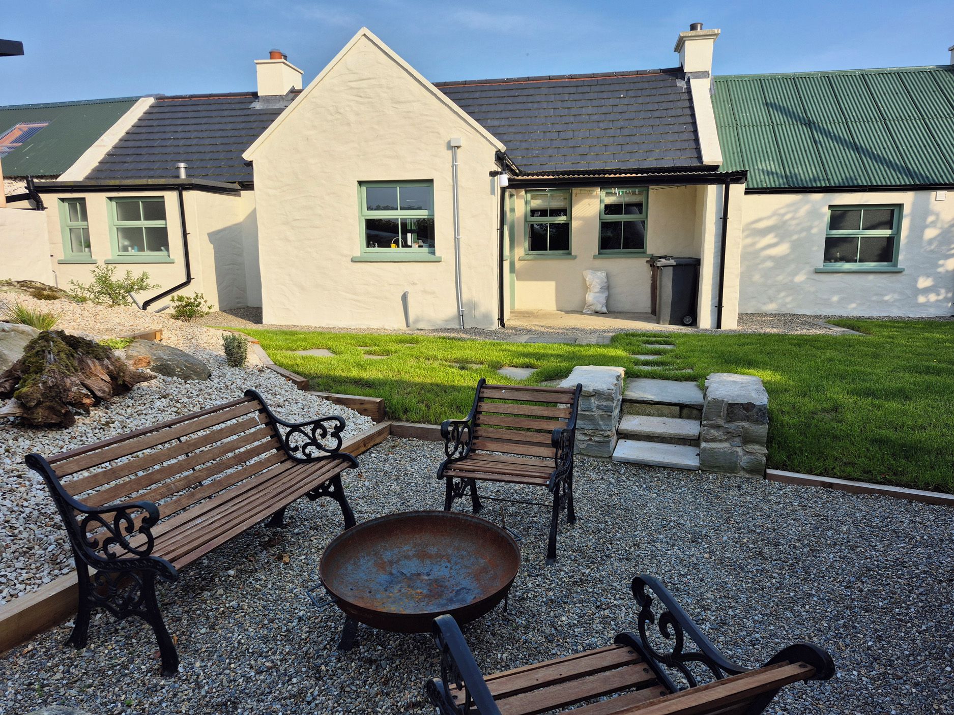 A house with a gravel patio, two wooden benches, a metal table, and a green lawn.