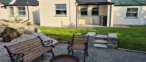 A house with a gravel patio, two wooden benches, a metal table, and a green lawn.
