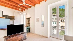 Un vaste espace de vie doté d’un plafond en bois, d’un téléviseur à écran plat posé sur un meuble en bois, d’un balcon offrant une vue sur les bâtiments et la verdure, ainsi que d’un coin cuisine équipé d’une cuisinière et de placards.