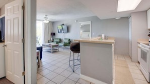 A modern kitchen with a central island, a dining area with green chairs, and a living room with a TV and a ceiling fan. - The Barbados | Private Balcony, Pool View! (Key West)
