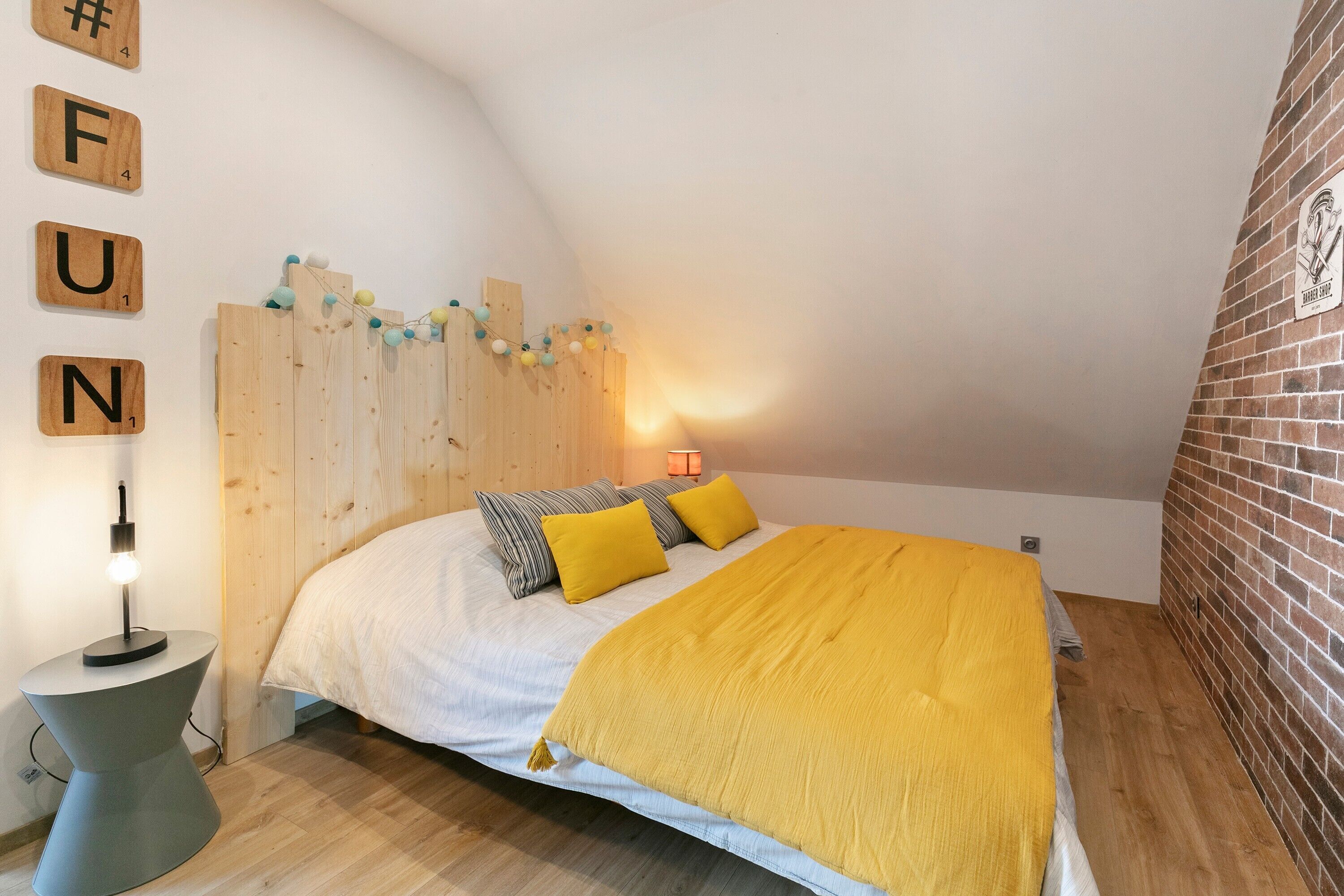 Une chambre avec un plafond en pente, un lit recouvert d’une couverture jaune, une tête de lit en bois et une lampe fixée au mur.