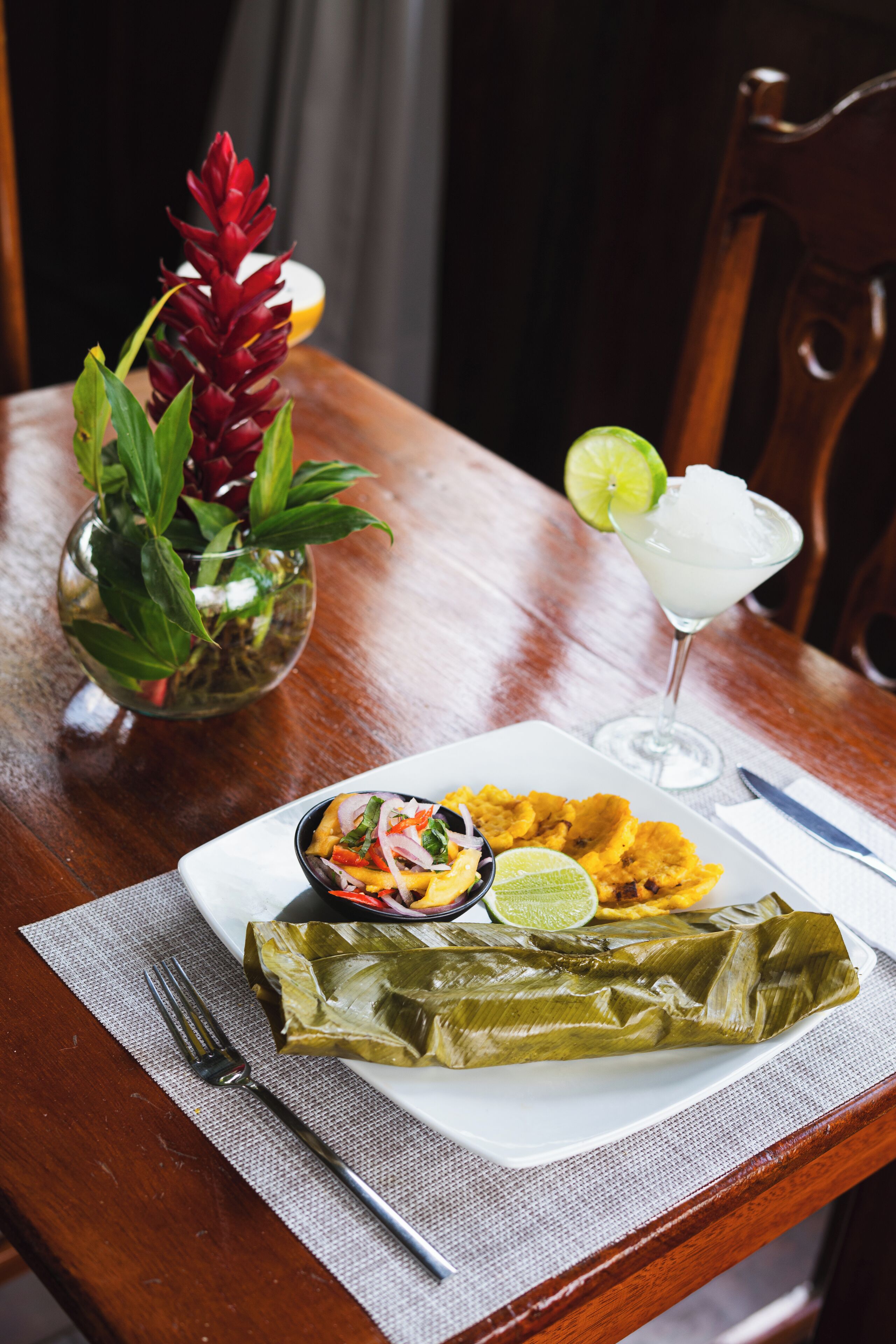 A plate of food including a tamale, fried plantains, and a lime drink, with a fork and knife on a table.