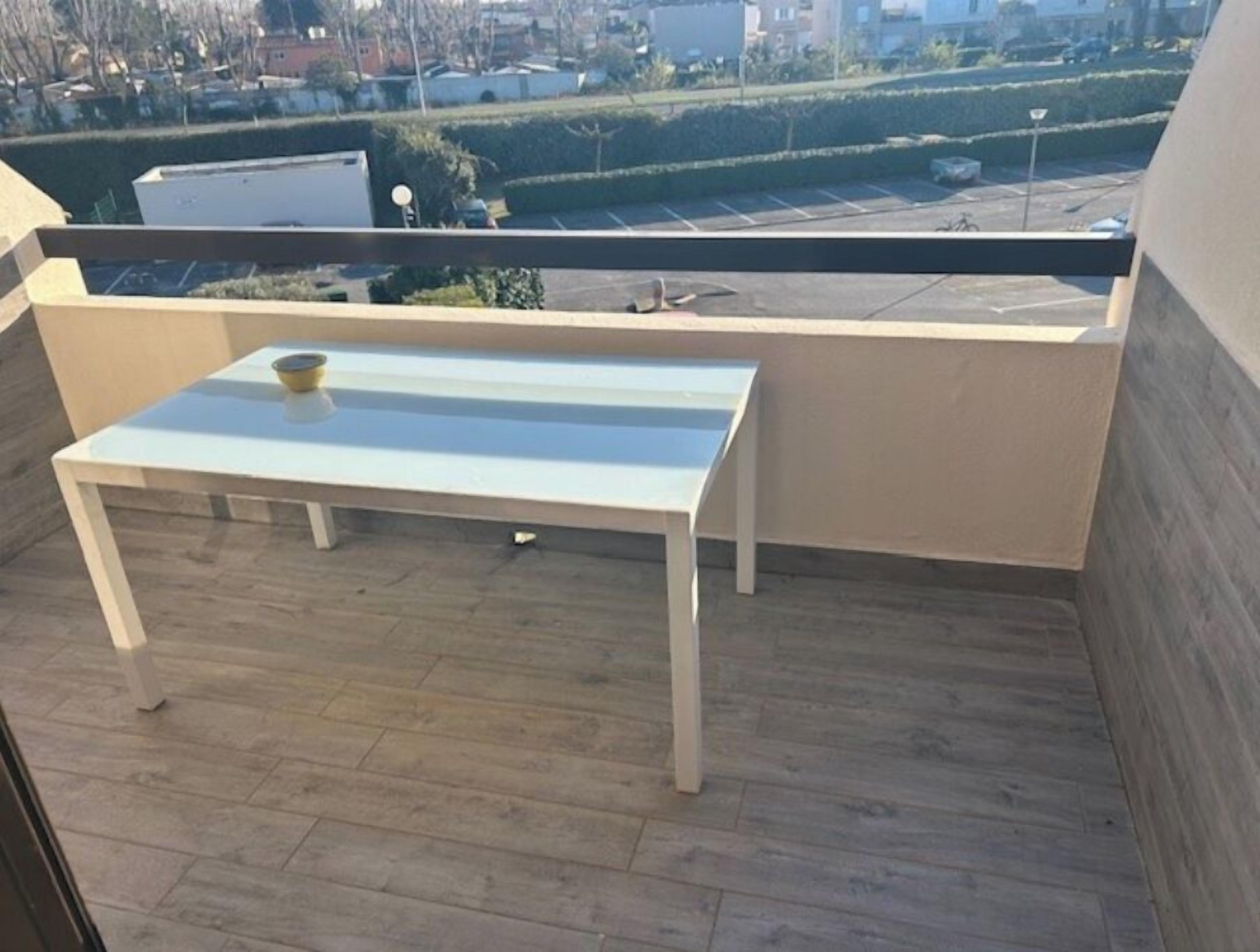A balcony with a white table and a bowl, overlooking a parking area and buildings.