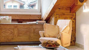 Une pièce chaleureuse avec un plafond en bois, une chaise blanche, une table sur laquelle se trouve un saladier rempli de fruits, et une fenêtre donnant sur des maisons.