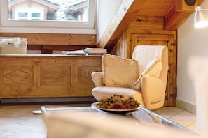Une pièce chaleureuse avec un plafond en bois, une chaise blanche, une table sur laquelle se trouve un saladier rempli de fruits, et une fenêtre donnant sur des maisons.