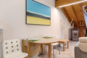 A dining area with a table set for two, a white chair, and a painting of a beach scene on the wall. - Apartment 'Il Nido' with Mountain View, Wi-Fi and Air Conditioning (Aosta)