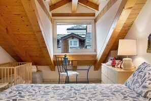 A bedroom with a sloped ceiling, a window with a view of a house, a bed with patterned bedding, a desk with a lamp, and a chair. - Apartment 'Il Nido' with Mountain View, Wi-Fi and Air Conditioning (Aosta)