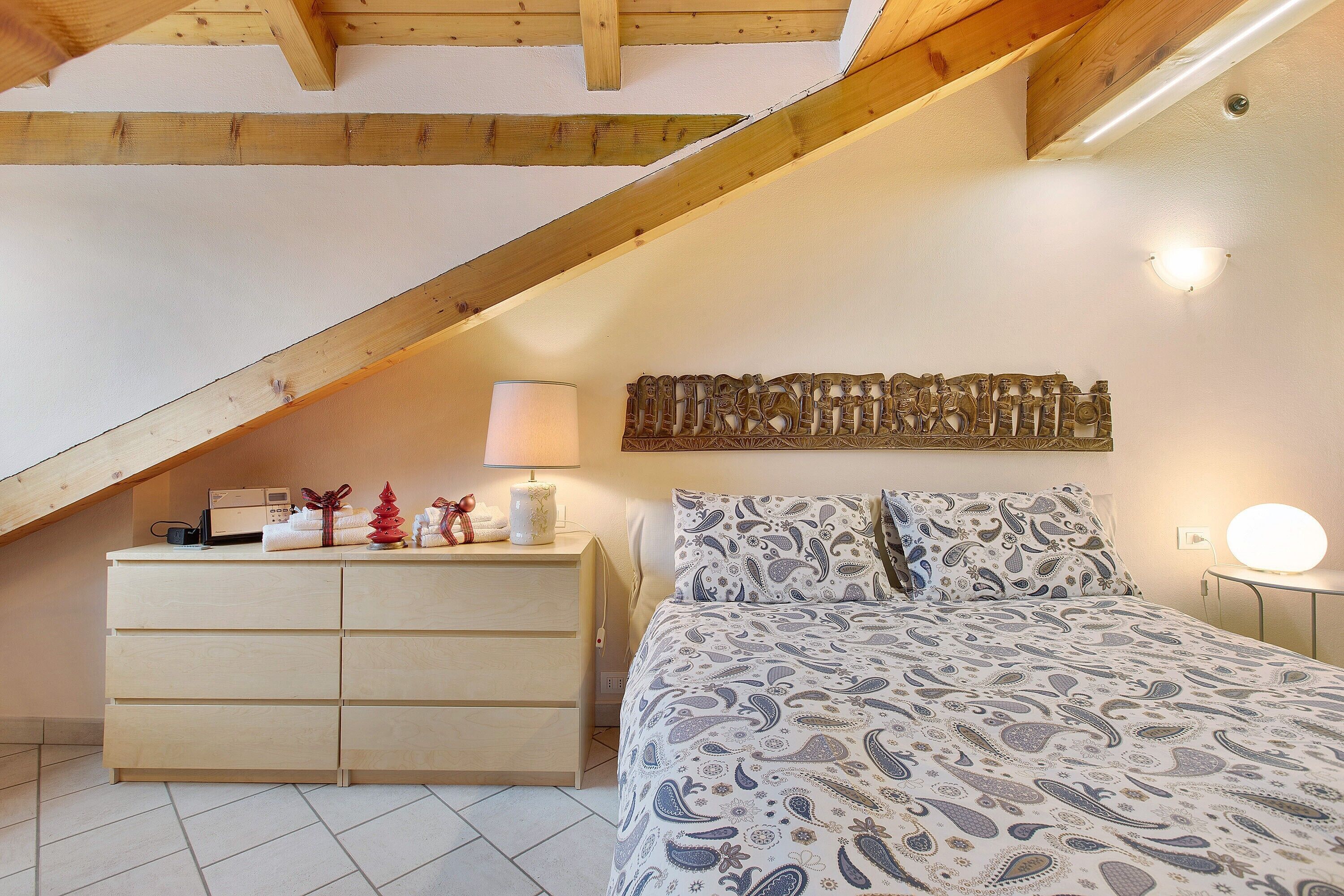 Ein Schlafzimmer mit einem Bett, einer Kommode, einer Wandleuchte und einer Decke mit freiliegenden Holzbalken.
