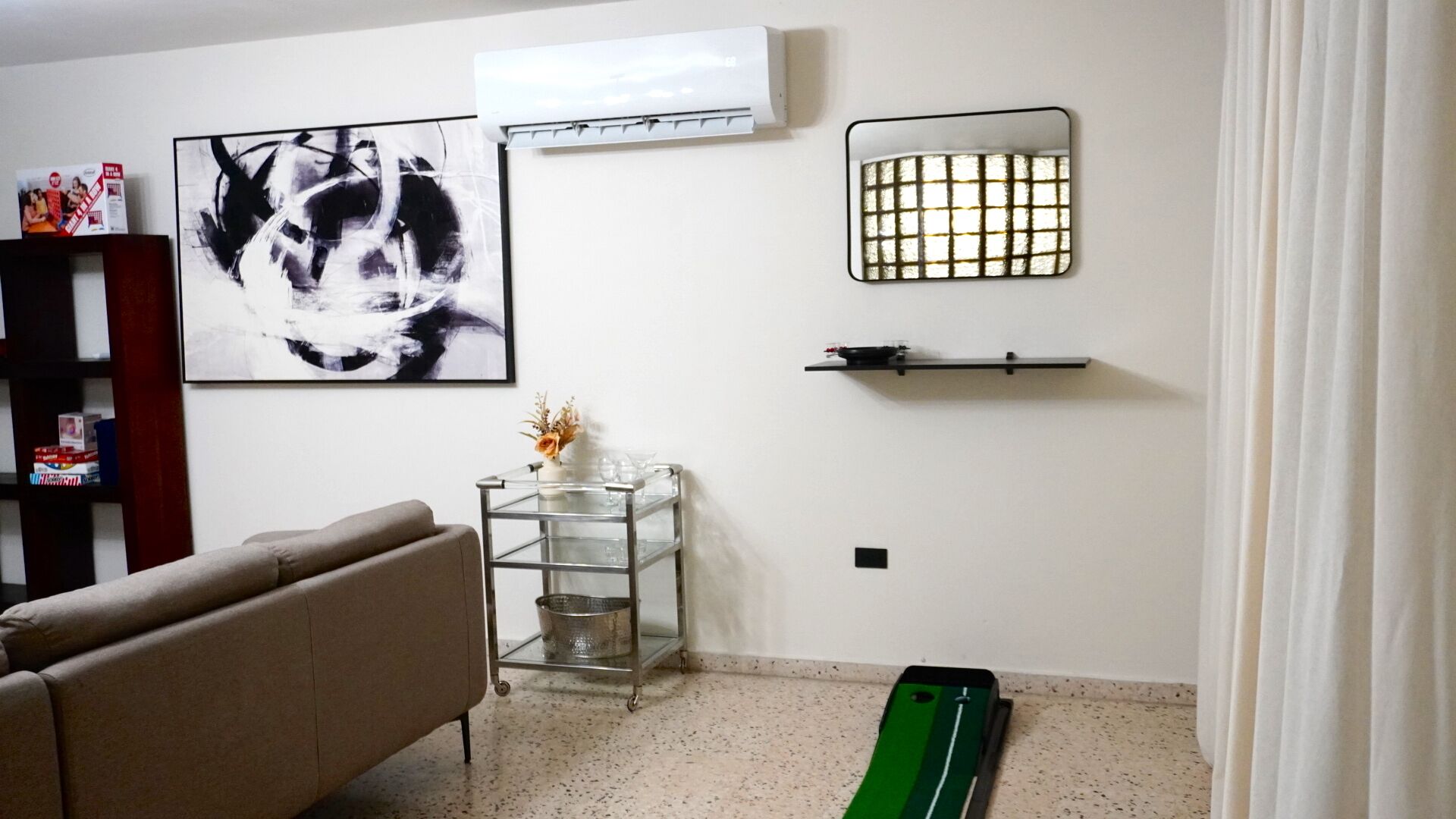 A living room with a sofa, a glass table with a vase, a black and white painting, an air conditioner, and a green object on the floor.