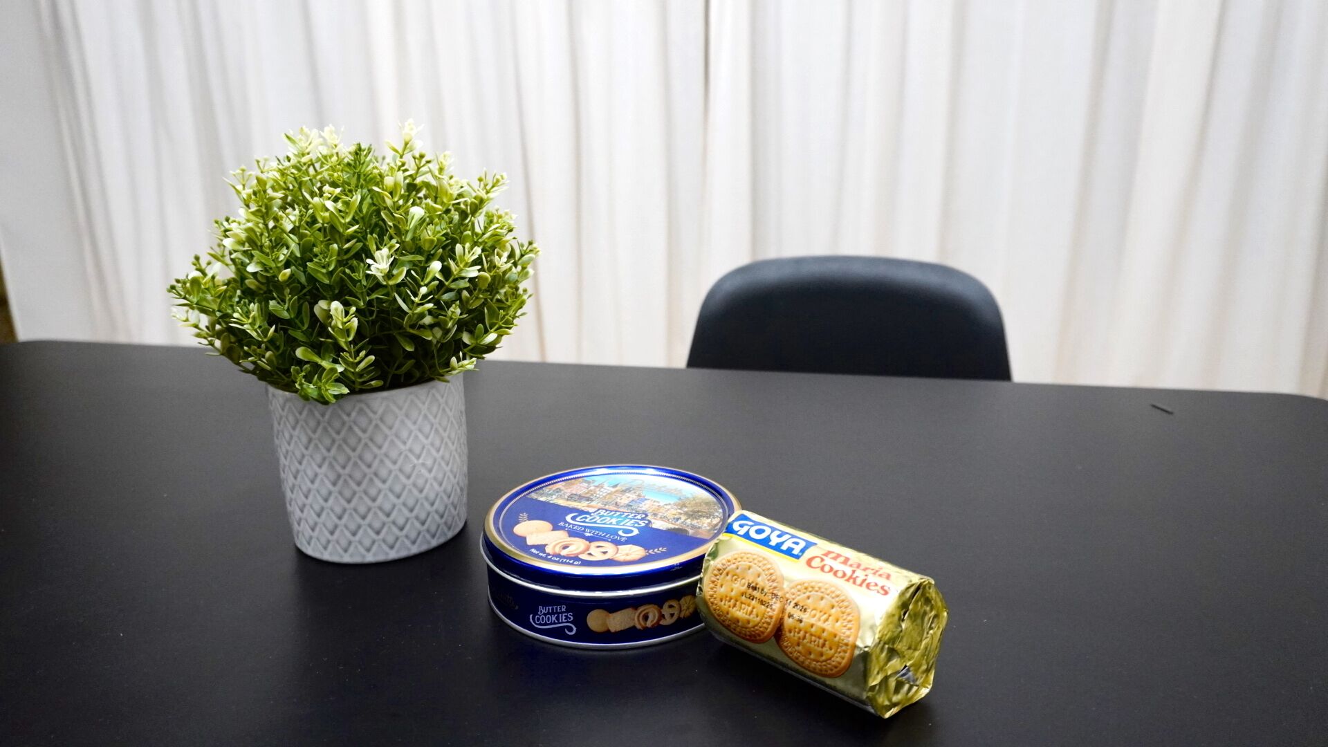 A small potted plant, a tin of butter cookies, and a partially unwrapped pack of Maria Cookies on a table.