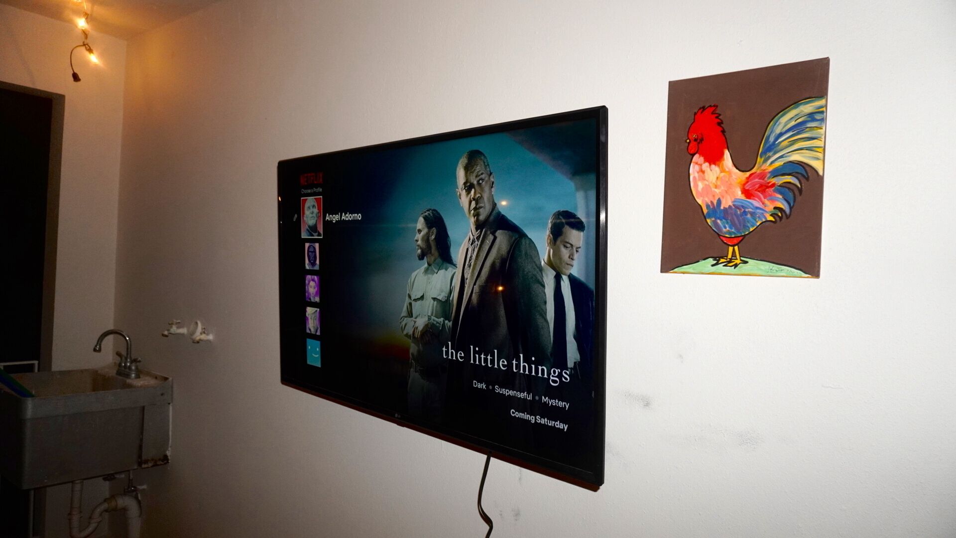 Et fjernsyn ophængt på en væg, der viser en filmplakat for 'The Little Things', med et billede af en hanekylling ved siden af.