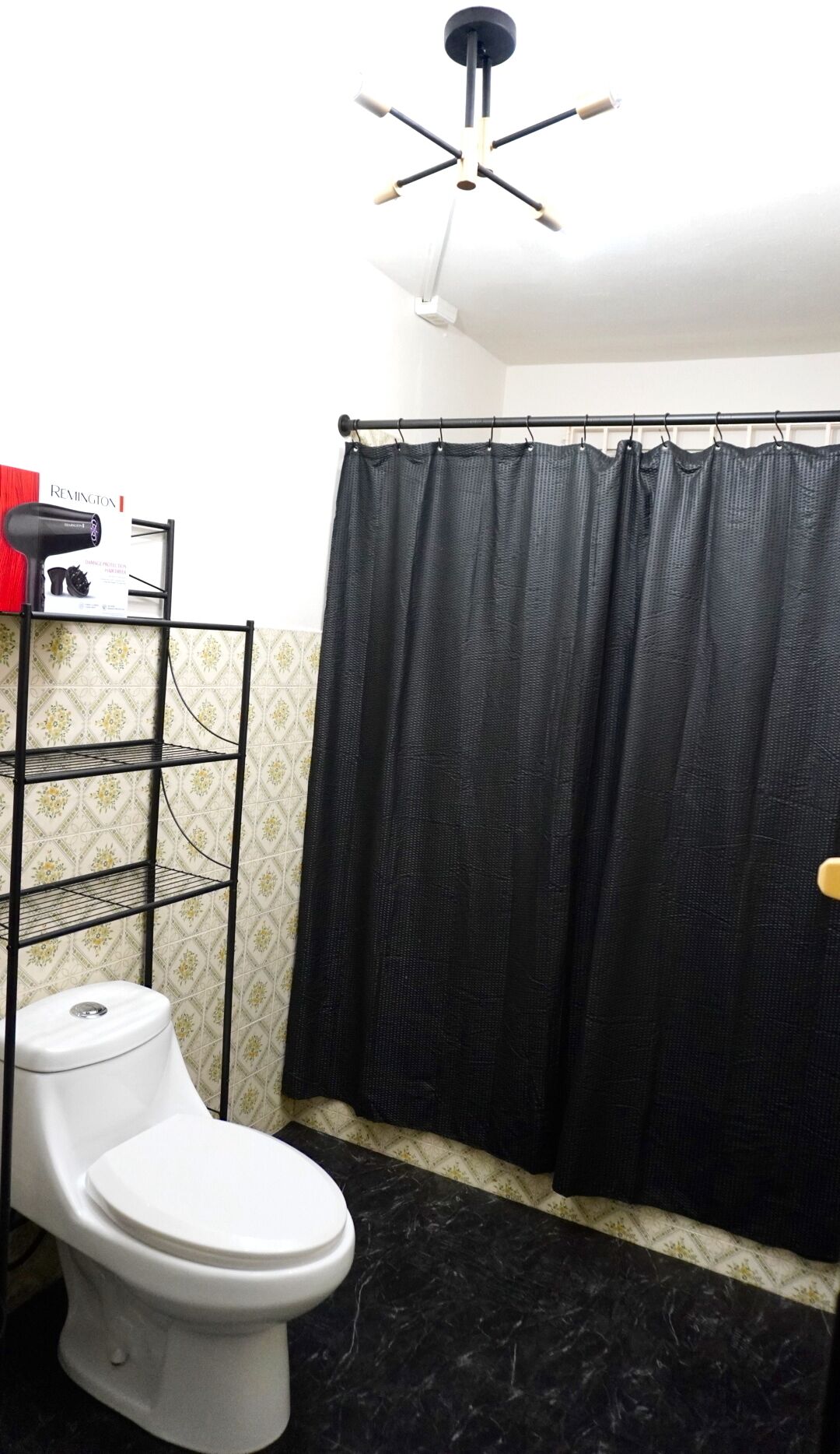 A bathroom with a toilet, a black shower curtain, a ceiling fan, and a shelf with toiletries.