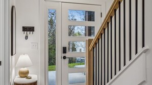 Un interior moderno con una escalera de madera, una puerta blanca con paneles de vidrio y una mesita redonda de madera con una lámpara.
