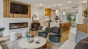 Una sala de estar moderna con chimenea, asientos cómodos, un comedor y una cocina con armarios de madera e iluminación colgante.