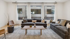 A modern living room with a sectional sofa, a coffee table, and a side table. There are large windows with curtains, a vase with branches, and a lamp on the side table. - NEW Modern Nashville Escape with Rooftop Hot Tub (Nashville)