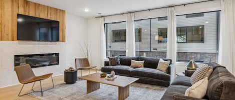 A modern living room with a large flat-screen TV mounted on a wooden panel, a fireplace, a sectional sofa, and a coffee table.