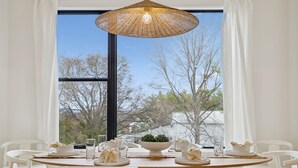 A dining area with a round table, white chairs, a woven pendant light, and a view of trees through large windows. - Luxury Nash Home w/ Rooftop Downtown Skyline Views (Nashville)