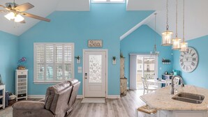 A spacious room with a high ceiling, a ceiling fan, a large window with shutters, a door with a frosted glass panel, a kitchen area with a sink and countertop, and a dining area with a table and chairs. - Molly's Beachside Cottage 3BR 2BA Almost Oceanfront in Atlantic Beach (Atlantic Beach)