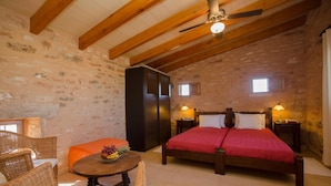 Une chambre à coucher rustique avec un plafond en bois, un lit recouvert d’une courtepointe rouge, des meubles en osier et une petite table sur laquelle sont disposés des fruits.