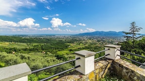Vista panoramica da un balcone con ringhiera in pietra, che si affaccia su una valle rigogliosa e montagne lontane sotto un cielo azzurro limpido.