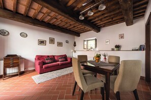 Una sala de estar acogedora con un sofá rojo, una mesa de comedor de madera y sillas. Hay luces fijadas a la pared, una ventana y un estante con libros y un jarrón de flores.