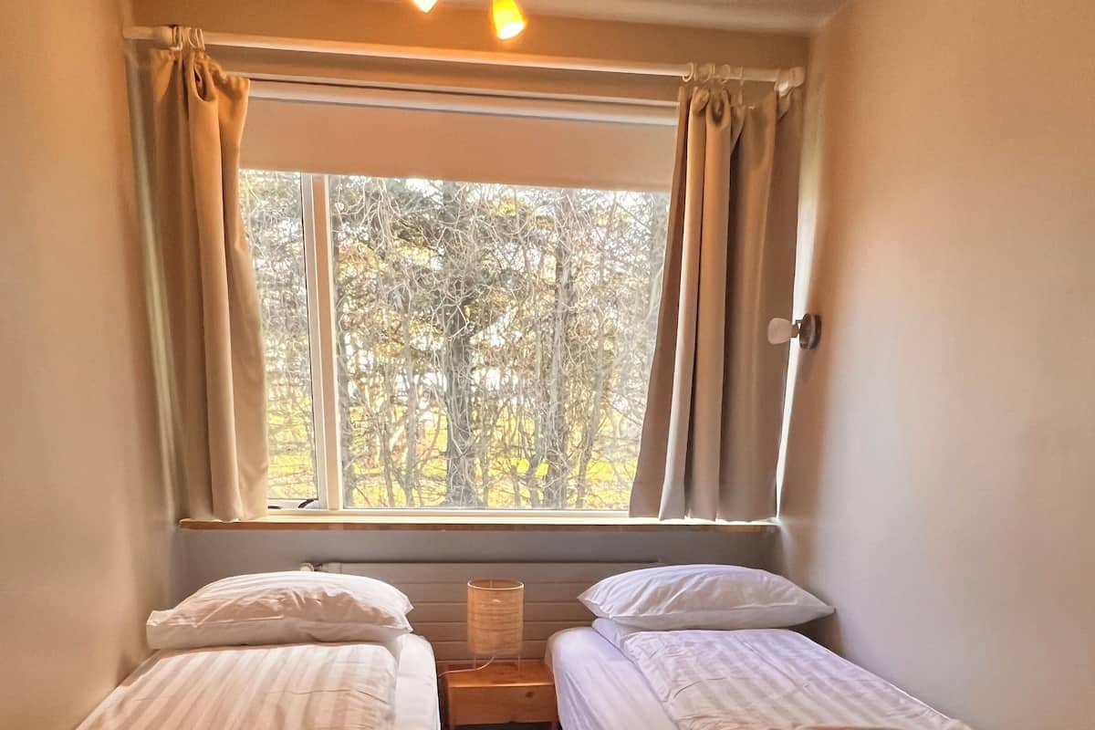 A room with two single beds, a nightstand, a lamp, and a window with curtains.