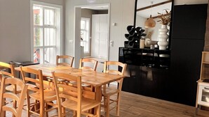 A dining area with a wooden table and chairs, a woven ceiling light, and a kitchen area with black cabinets and a mirror. - Kot Guesthouse (Hvolsvöllur)