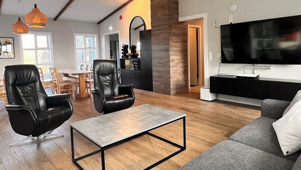 A spacious living room with a large gray sofa, two black leather chairs, a wooden coffee table, and a flat-screen TV mounted on the wall. - Kot Guesthouse (Hvolsvöllur)