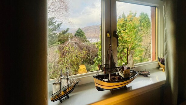 Uma janela com dois modelos de navios em um parapeito, com vista para uma paisagem com árvores e colinas.
