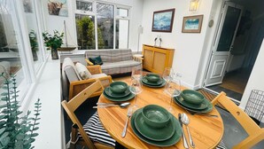 Uma área de refeições com uma mesa de madeira redonda posta para quatro pessoas, pratos verdes e talheres. Há um sofá e um armário ao fundo.