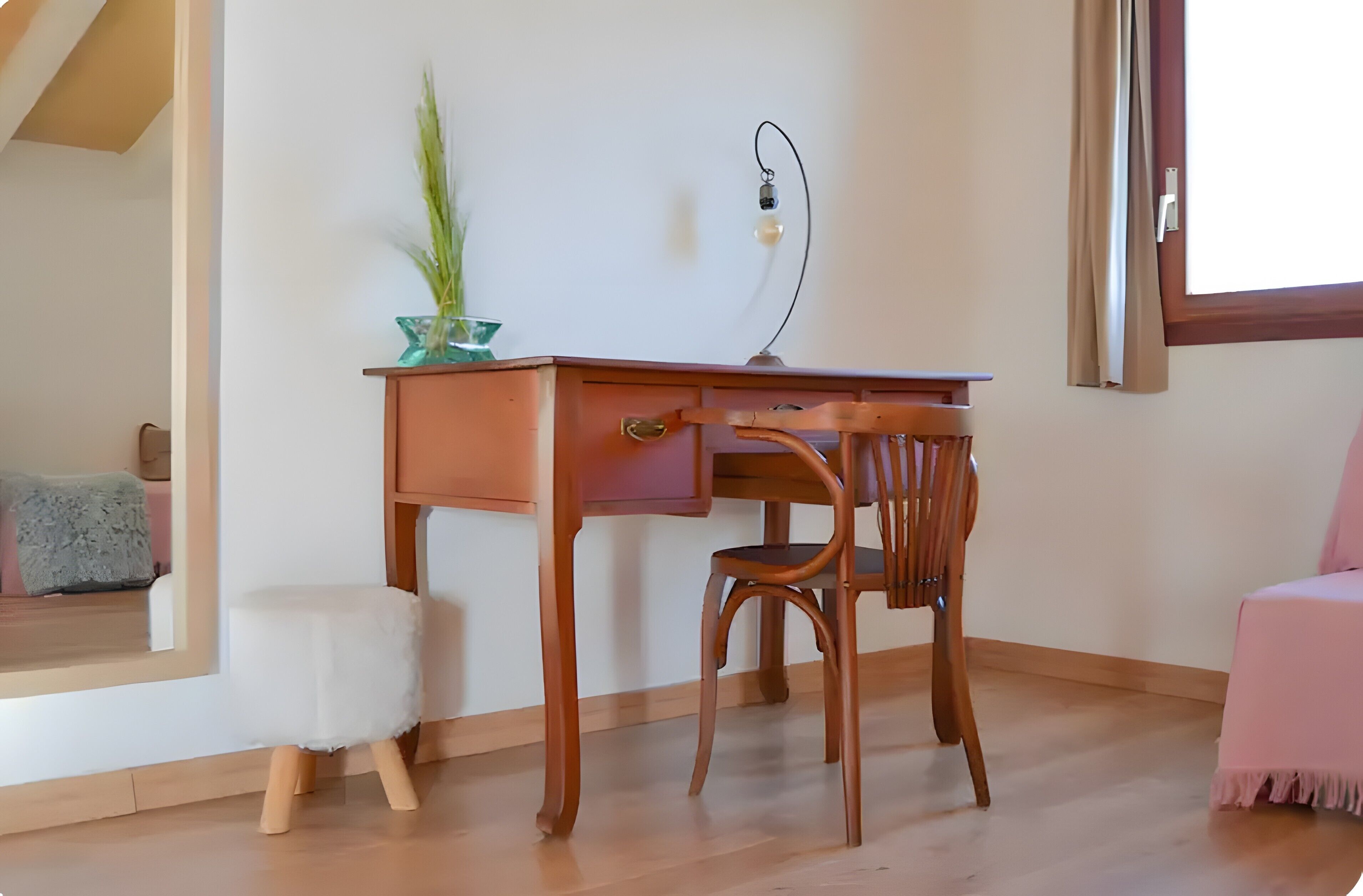 Un escritorio de madera con un cajón, una silla, un taburete pequeño, una planta en maceta y una ventana con cortinas.
